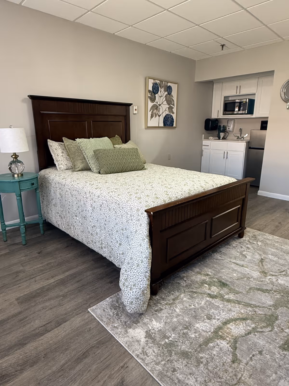 A bedroom with a wooden bed frame and a bed made with a white and green patterned bedspread and multiple green pillows. To the left of the bed is a small teal nightstand with a lamp. On the wall above the bed is a framed artwork featuring blue flowers. In the background, there is a kitchenette area with white cabinets, a microwave, a sink, and a small refrigerator. The floor is wood with a light-colored patterned rug partially under the bed.