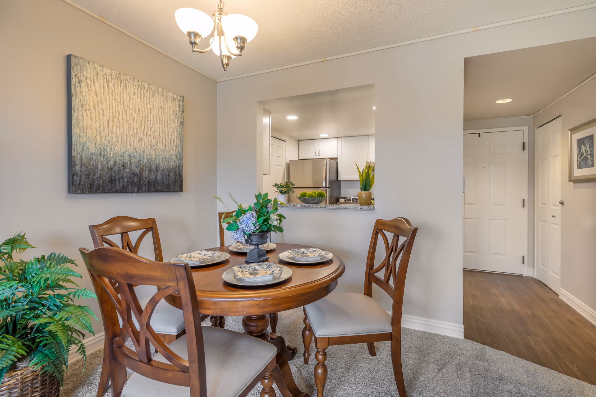 A dining area with a round wooden table set for four with plates and napkins. Four wooden chairs with cushioned seats surround the table. A small floral centerpiece is on the table. The dining area has beige walls, a hanging light fixture, and a large abstract painting on the wall. Through a pass-through window, a kitchen with white cabinets and a stainless steel refrigerator is visible. To the right, there is a hallway with white doors and wood flooring.