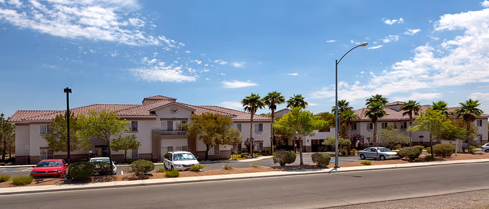 Street view of a two-story beige assisted living building with red tile roofs, palm trees, landscaping, and parked cars along the front.