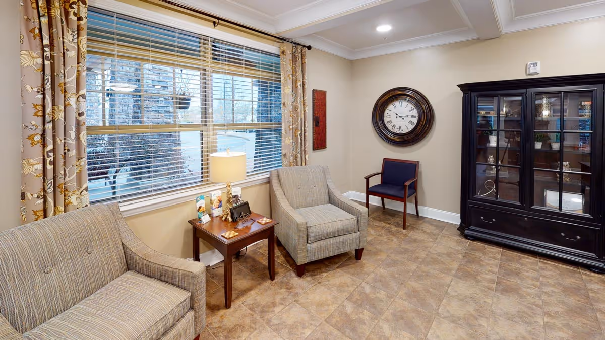 A cozy sitting area in Oakview Park featuring a plaid upholstered loveseat and armchair, a wooden side table with a lamp and brochures, a large window with floral curtains, a wall clock, a wooden chair with blue upholstery, and a dark wooden cabinet with glass doors displaying decorative items.