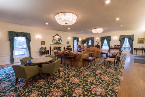 A spacious living room with floral patterned carpet, multiple seating areas including green armchairs around a round table, striped sofas, and wooden chairs. The room features large windows with green curtains, two elegant chandeliers, a fireplace with a mirror above it, and various framed pictures on the walls.