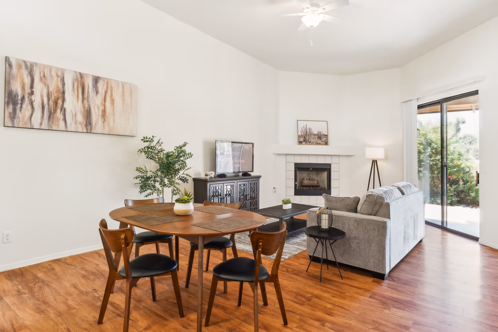 A bright living and dining area with wooden flooring. The dining area has a round wooden table with four chairs and placemats. The living area features a gray sofa, a black coffee table, a side table with a decorative lantern, a floor lamp, and a fireplace with a framed picture of cacti above it. A TV sits on a dark cabinet against the wall, and a large sliding glass door opens to a garden outside. A large abstract painting hangs on the wall above the dining table.