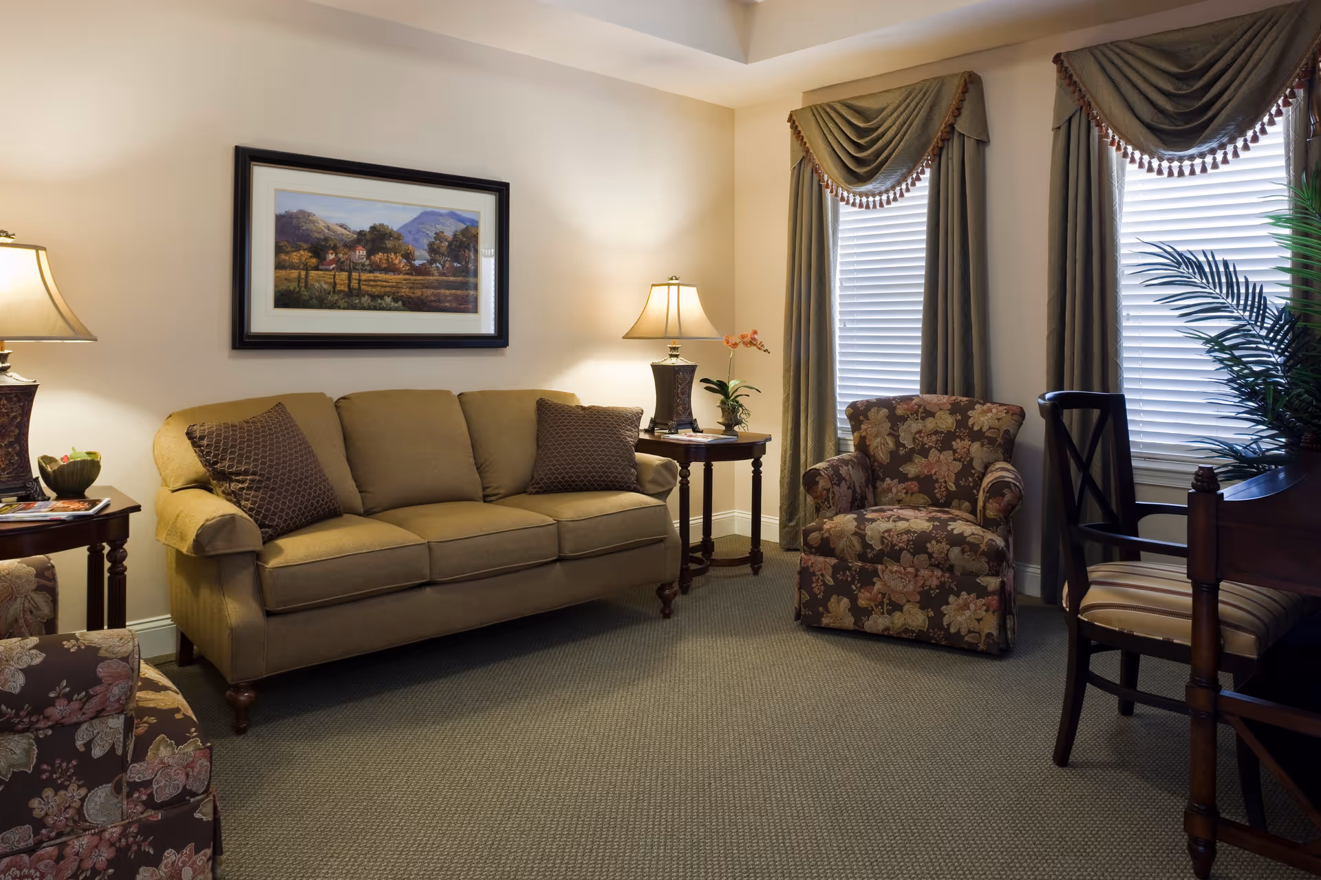 A cozy living room with a beige sofa, two floral armchairs, a wooden chair with striped cushion, two side tables with lamps, a framed landscape painting on the wall, and two windows with green drapes and white blinds.