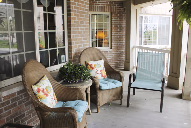 Covered porch with wicker chairs, a small table holding a potted plant, and a metal chair in front of brick walls and windows.
