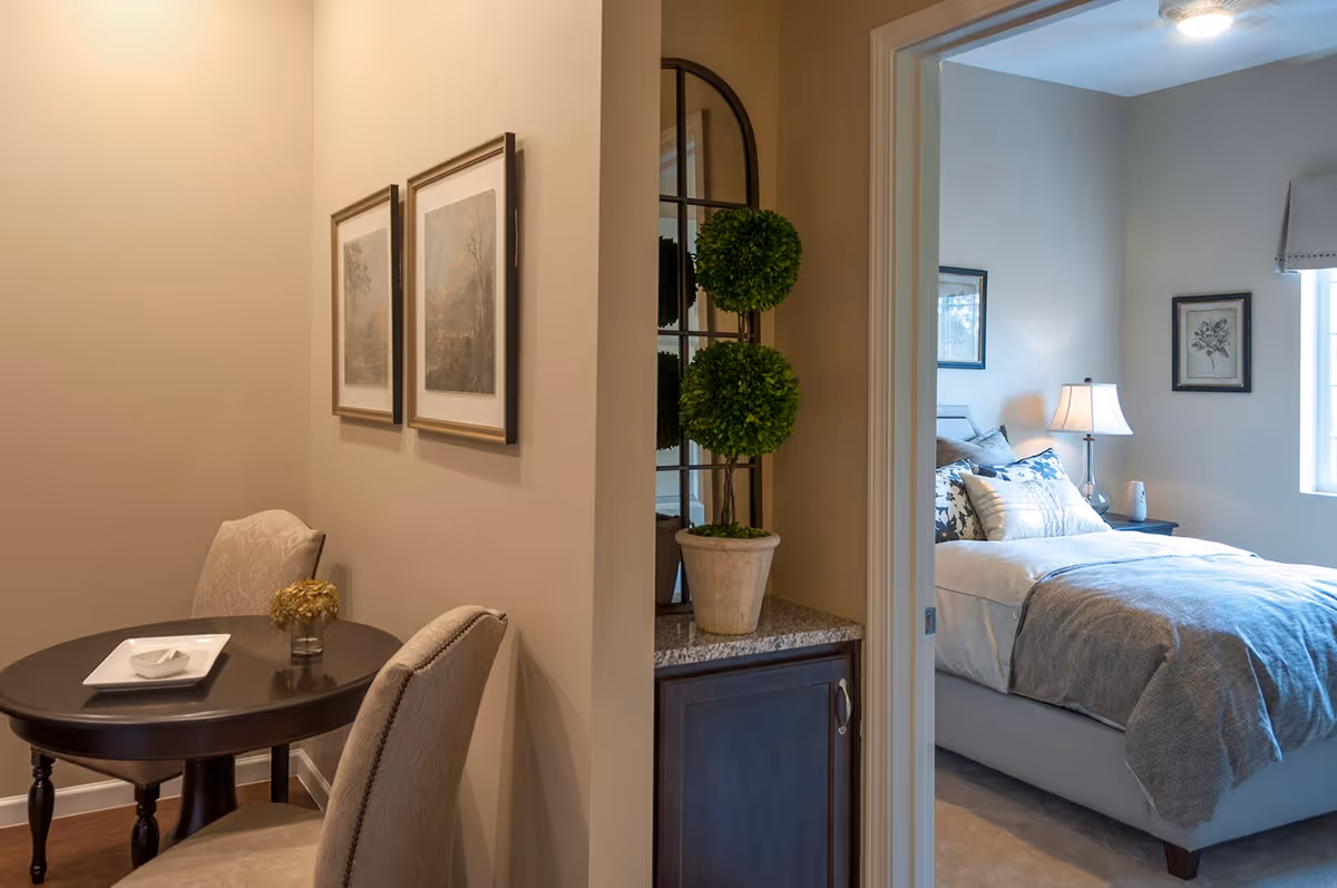 A cozy interior space showing a small round dark wood table with two upholstered chairs, a small vase with dried flowers, and two framed pictures on the wall. To the right, an open doorway reveals a bedroom with a neatly made bed, decorative pillows, a bedside table with a lamp, and framed artwork on the walls.