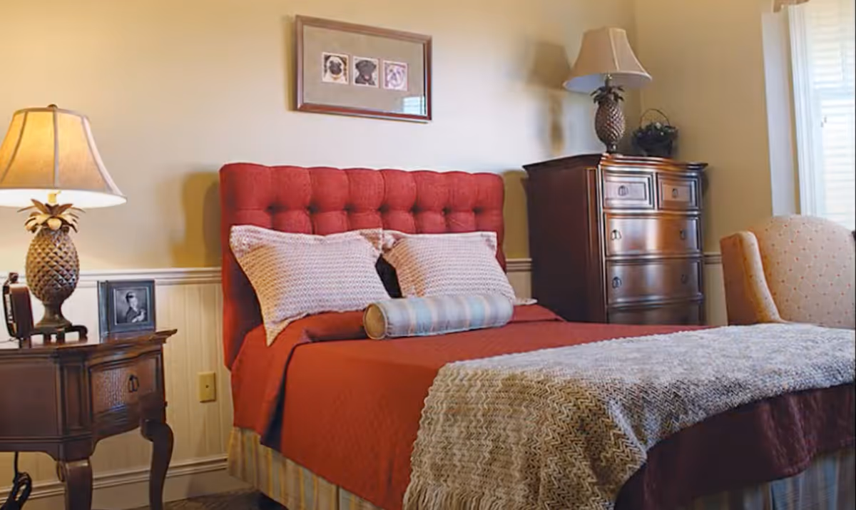 Cozy bedroom with a red tufted headboard, pillows and throw blanket, bedside tables with lamps, a dresser and an armchair.