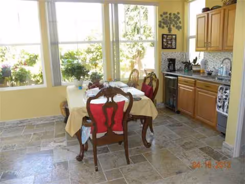A dining area and kitchenette with a wooden table, red chairs, and sunlight from large windows.