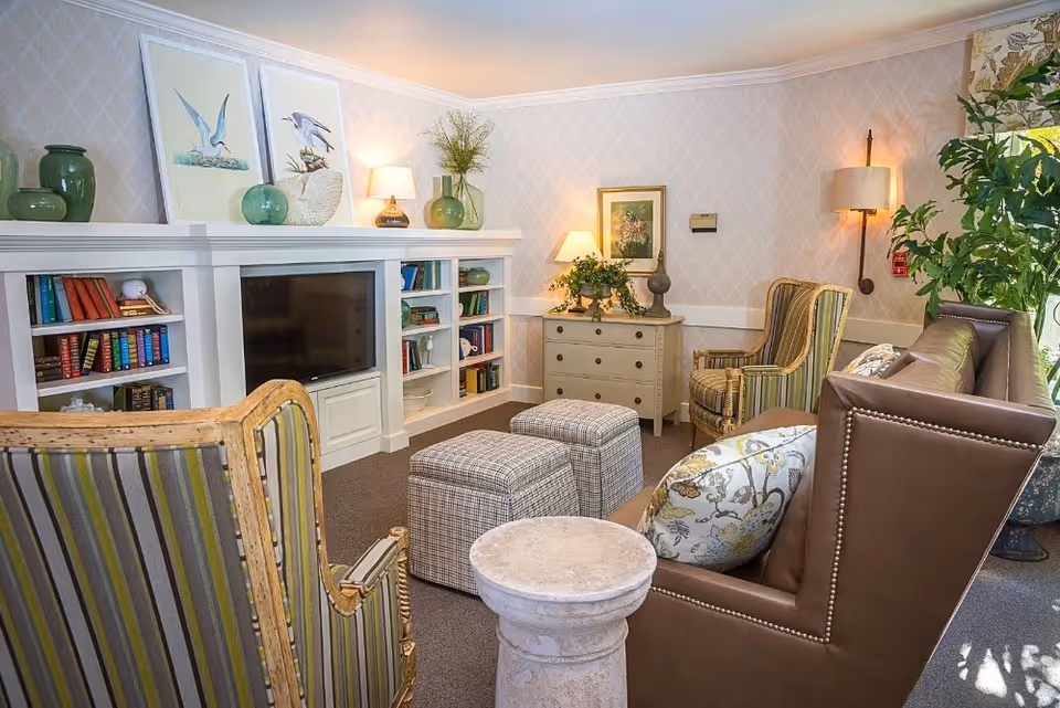 A cozy living room with a brown leather sofa adorned with patterned pillows, two striped armchairs, and two plaid ottomans. A white built-in bookshelf with books and decorative items surrounds a flat-screen TV. The room is softly lit by table lamps and a wall sconce, with framed bird artwork and plants adding to the decor.