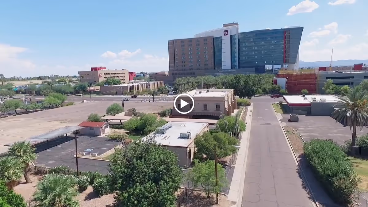 Aerial view of a suburban area with a road running through the center, surrounded by buildings, trees, and parking lots. In the background, there is a large multi-story hospital building with clear skies above.