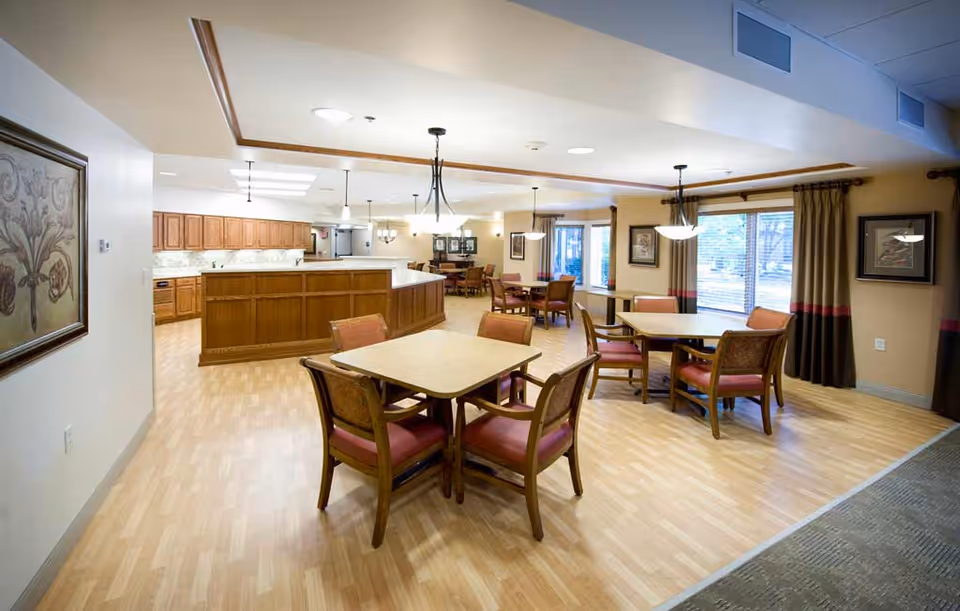 A spacious dining area in a retirement community featuring multiple square tables with four wooden chairs each, cushioned with red upholstery. The room has light wood flooring, beige walls, and large windows with brown and red curtains allowing natural light. The background shows a kitchen area with wooden cabinets and a long counter. Ceiling lights and hanging lamps illuminate the space, and framed artwork decorates the walls.