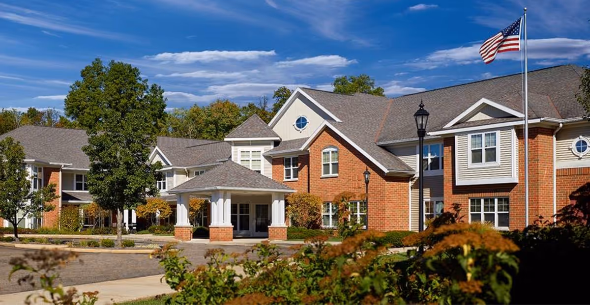 Front exterior of a two-story brick senior living building with a covered entrance, landscaping, and an American flag.
