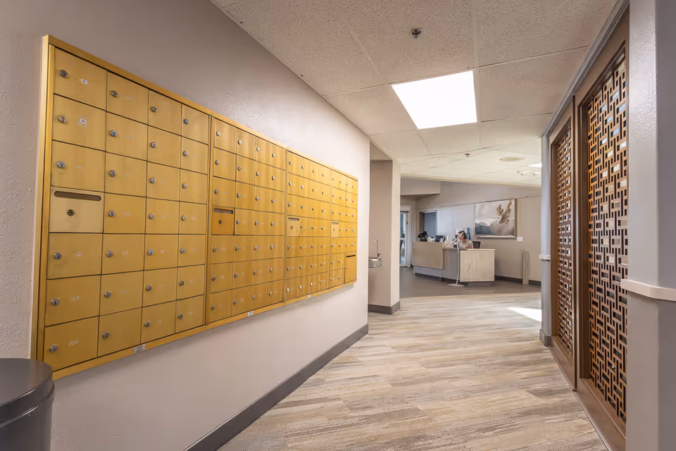 Interior hallway of a senior living facility with a wall-mounted bank of yellow mailboxes on the left and decorative wooden panels on the right. At the end of the hallway, there is a reception desk with a person sitting behind it. The floor is carpeted with a light pattern, and the ceiling has recessed lighting panels.