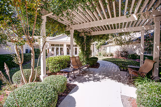 A serene garden pathway at Village Cove Assisted Living, featuring a pergola, comfortable seating, and well-maintained greenery.