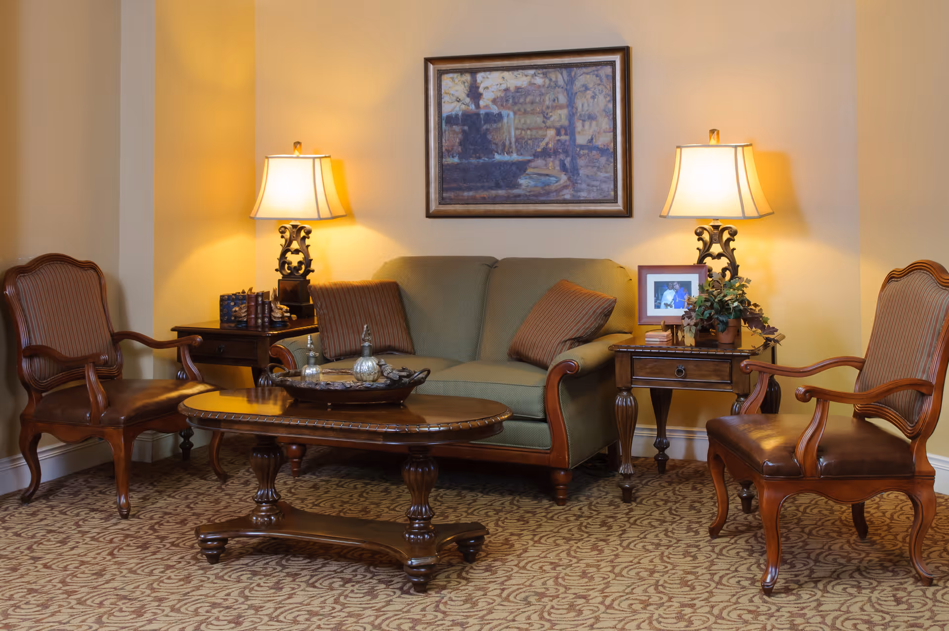 A cozy living room area with a green upholstered sofa flanked by two wooden side tables, each holding a decorative lamp. Two wooden armchairs with striped cushions face the sofa across a wooden coffee table with decorative items. A framed painting hangs on the wall above the sofa, and a framed photo and a potted plant are on the right side table. The room has patterned carpet and warm lighting.