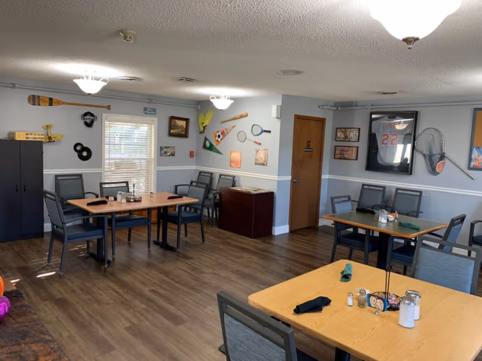 A dining room with several tables and chairs arranged neatly. The walls are decorated with sports memorabilia including paddles, a framed baseball jersey, a tennis racket, and various signs and pictures. The floor is wooden, and the room is well-lit with ceiling lights.