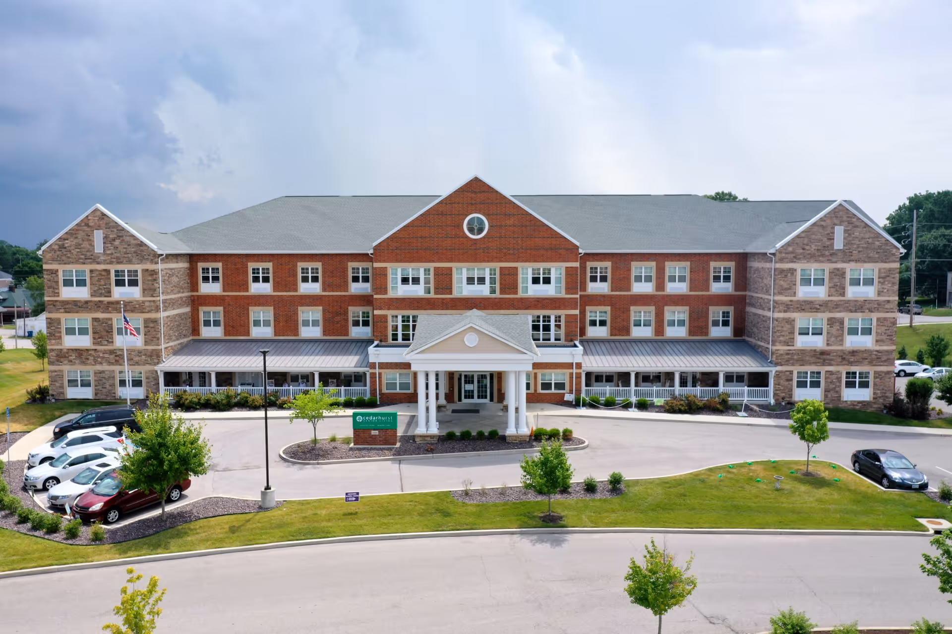 Front exterior view of Cedarhurst Senior Living of St. Charles, a large three-story building with a combination of brick and stone facade, multiple windows, a covered entrance with white columns, a circular driveway, and several parked cars. The surrounding area includes landscaped greenery and trees under a partly cloudy sky.