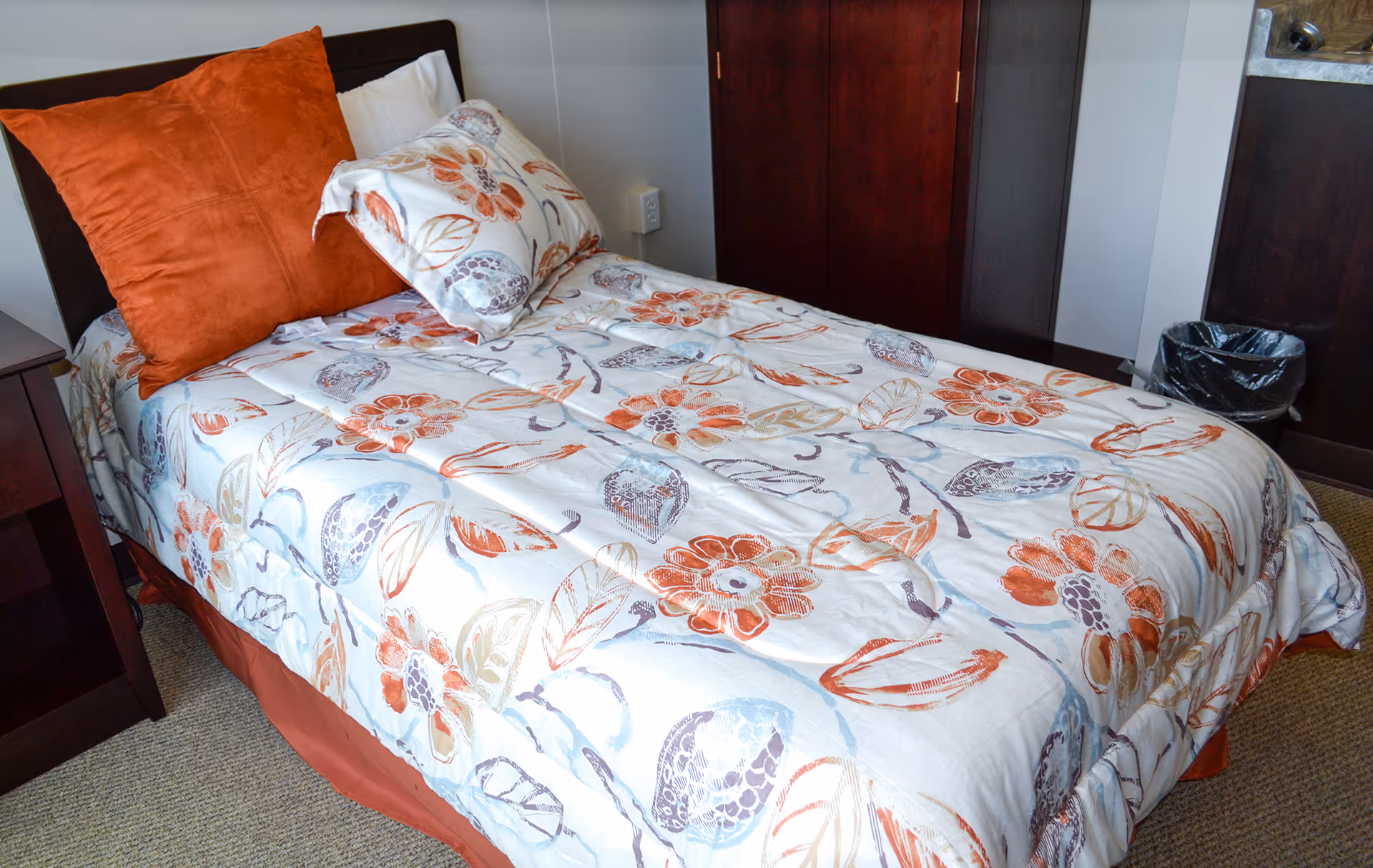Single bed with a floral bedspread and orange pillows in a neatly furnished bedroom with a nightstand and trash can.