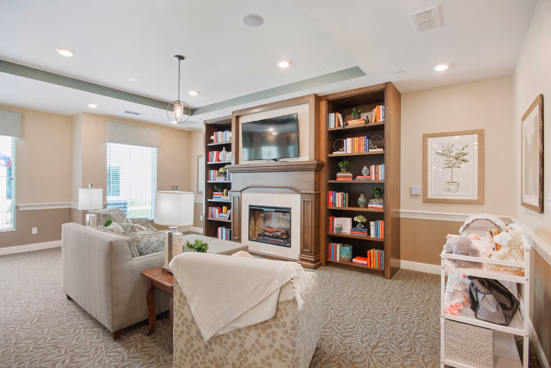 A cozy living room area with a beige sofa and armchair, a wooden coffee table, and two table lamps. The room features a built-in wooden bookshelf filled with books on either side of a fireplace with a mounted flat-screen TV above it. There are two large windows with blinds letting in natural light, and framed botanical artwork on the walls. A small white shelf with blankets and pillows is visible on the right side.
