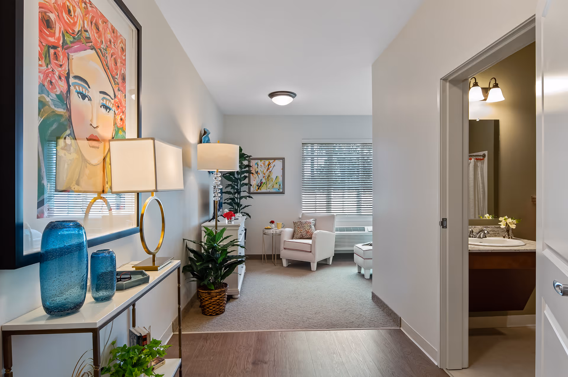 Interior view of a senior living facility room at The Vero at Bethlehem showing a hallway with a console table holding two blue glass vases and a lamp. The hallway leads to a sitting area with a white armchair, ottoman, side table, and a window with blinds. To the right, there is a bathroom entrance with a sink, mirror, and light fixture visible.