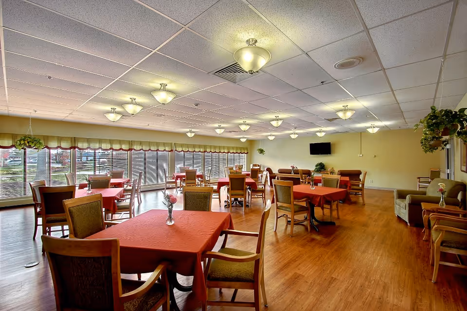 A spacious dining room with multiple tables covered in red tablecloths, each surrounded by wooden chairs with cushioned seats. The room has large windows with blinds allowing natural light to enter, and several ceiling lights providing additional illumination. There are plants hanging near the windows and a wall-mounted television on the far wall. The floor is wooden, and the overall atmosphere is warm and inviting.