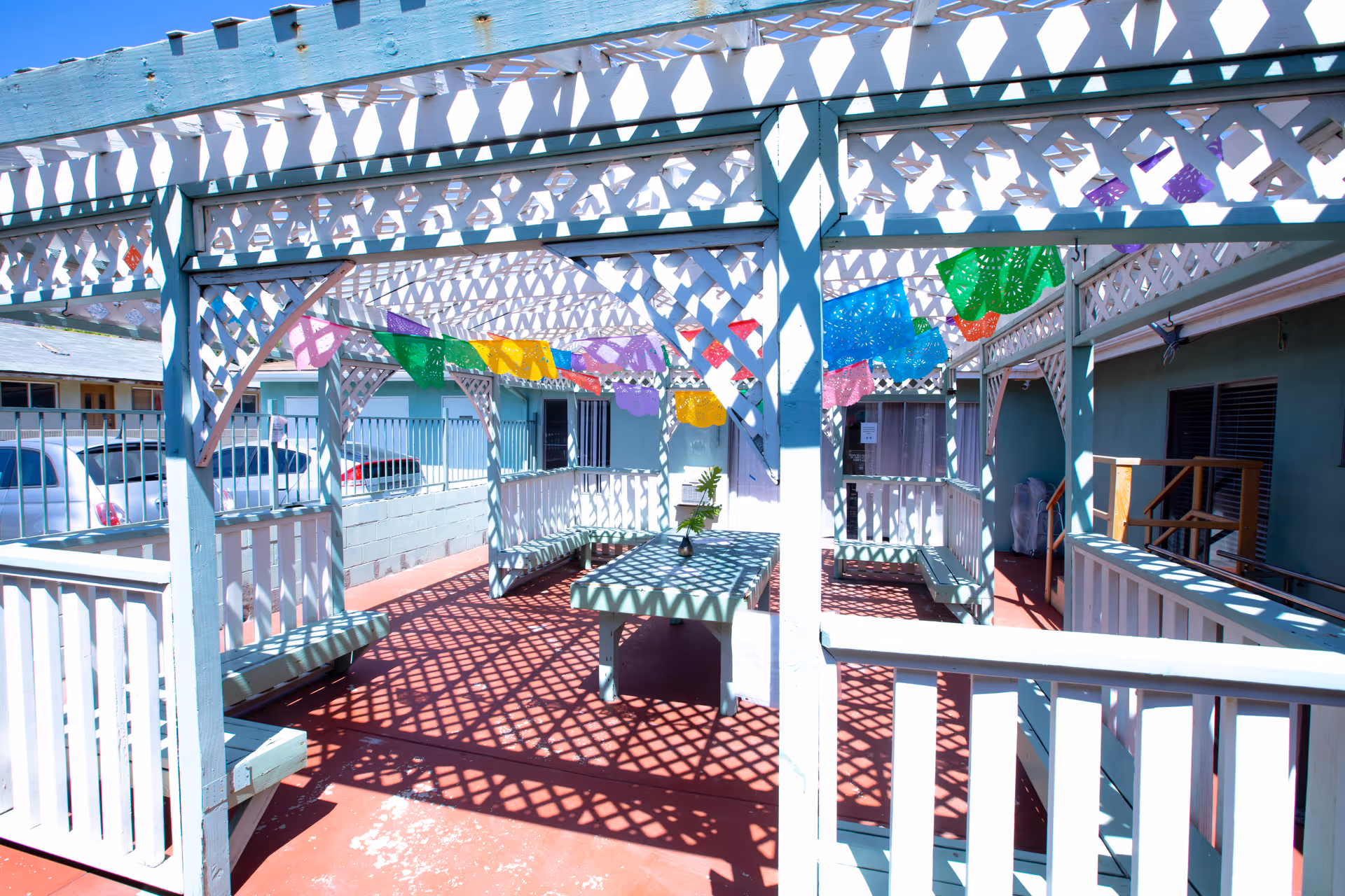 Outdoor patio area with white lattice pergola casting patterned shadows on the red floor. The space includes benches and a table with a small plant on it. Colorful decorative flags hang across the pergola. Surrounding buildings and parked cars are visible in the background.