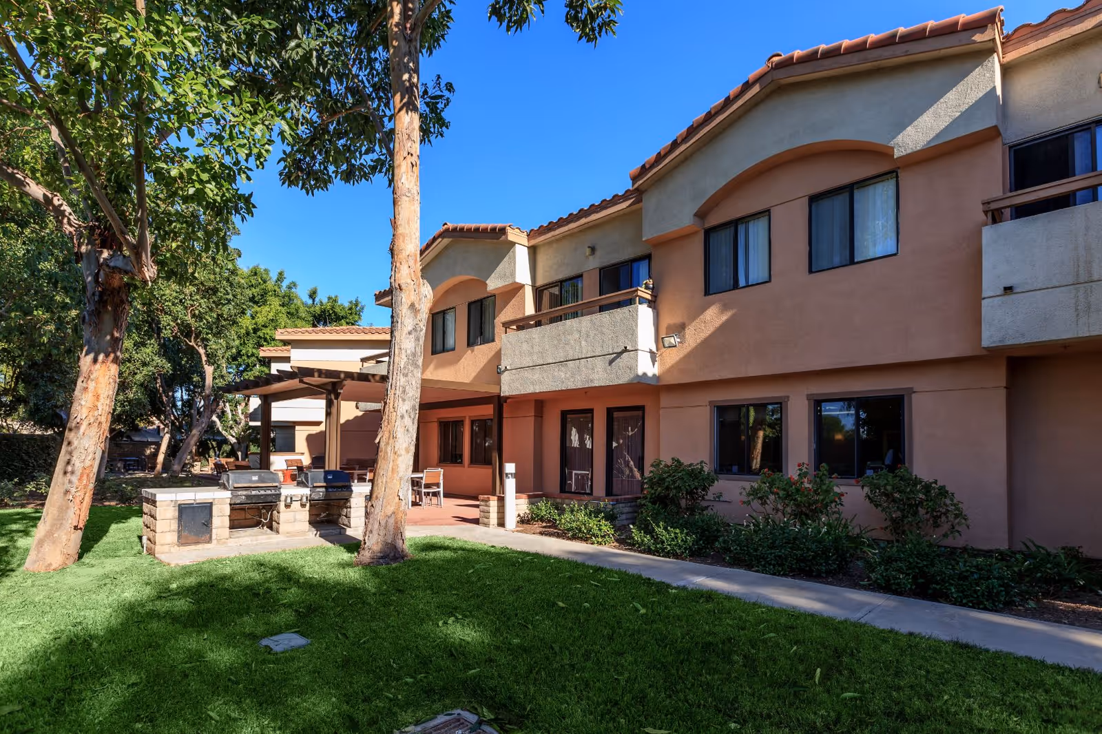 Outdoor view of a two-story residential building with beige and peach-colored walls, multiple windows, and balconies. In the foreground, there is a grassy lawn with trees and a built-in outdoor grill area with two grills and a countertop. The sky is clear and blue.