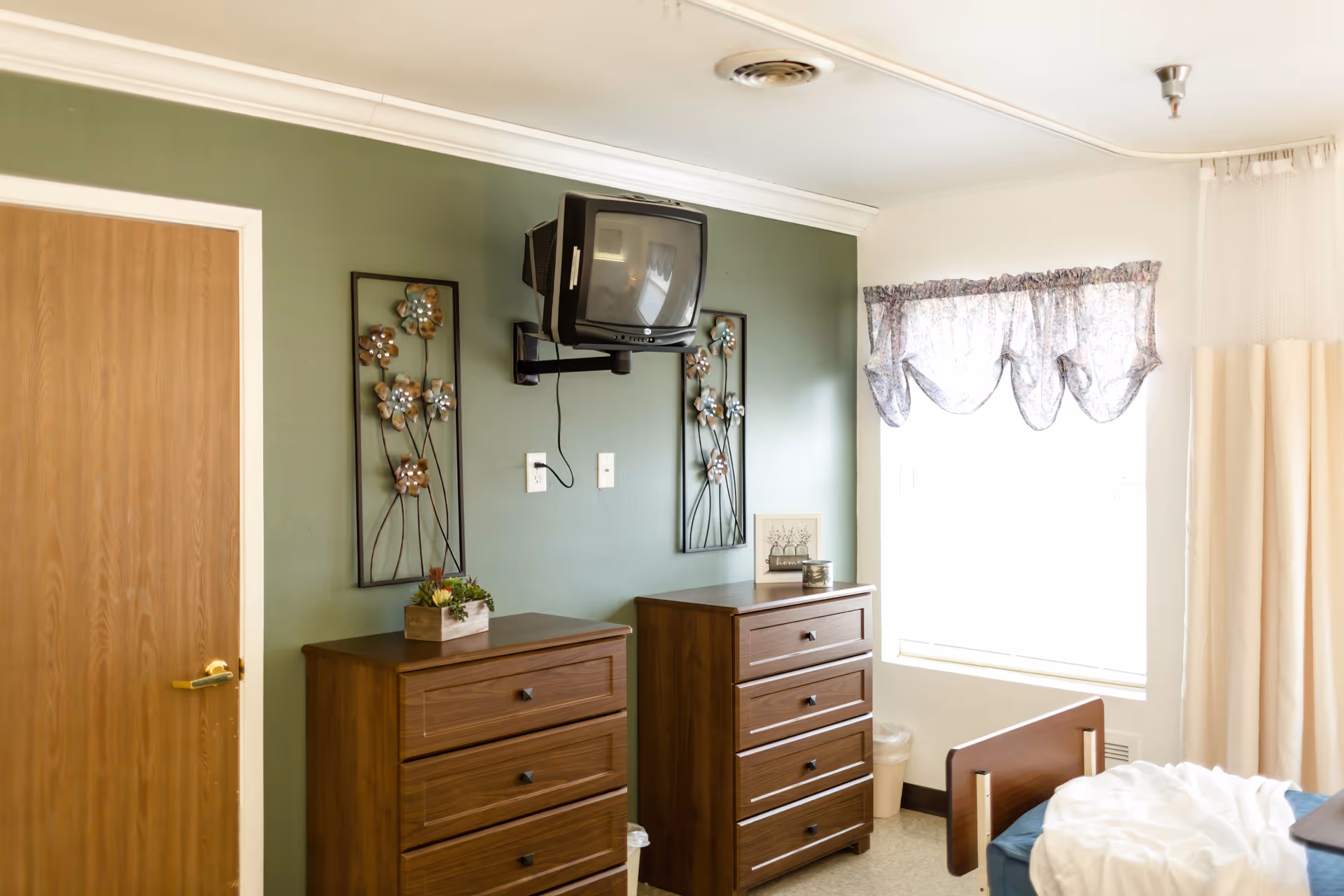 A room with two wooden dressers against a green wall, each decorated with small items. Above the dressers is a wall-mounted CRT television. To the right, there is a window with floral curtains and a bed partially visible with white bedding. A wooden door is on the left side of the image.