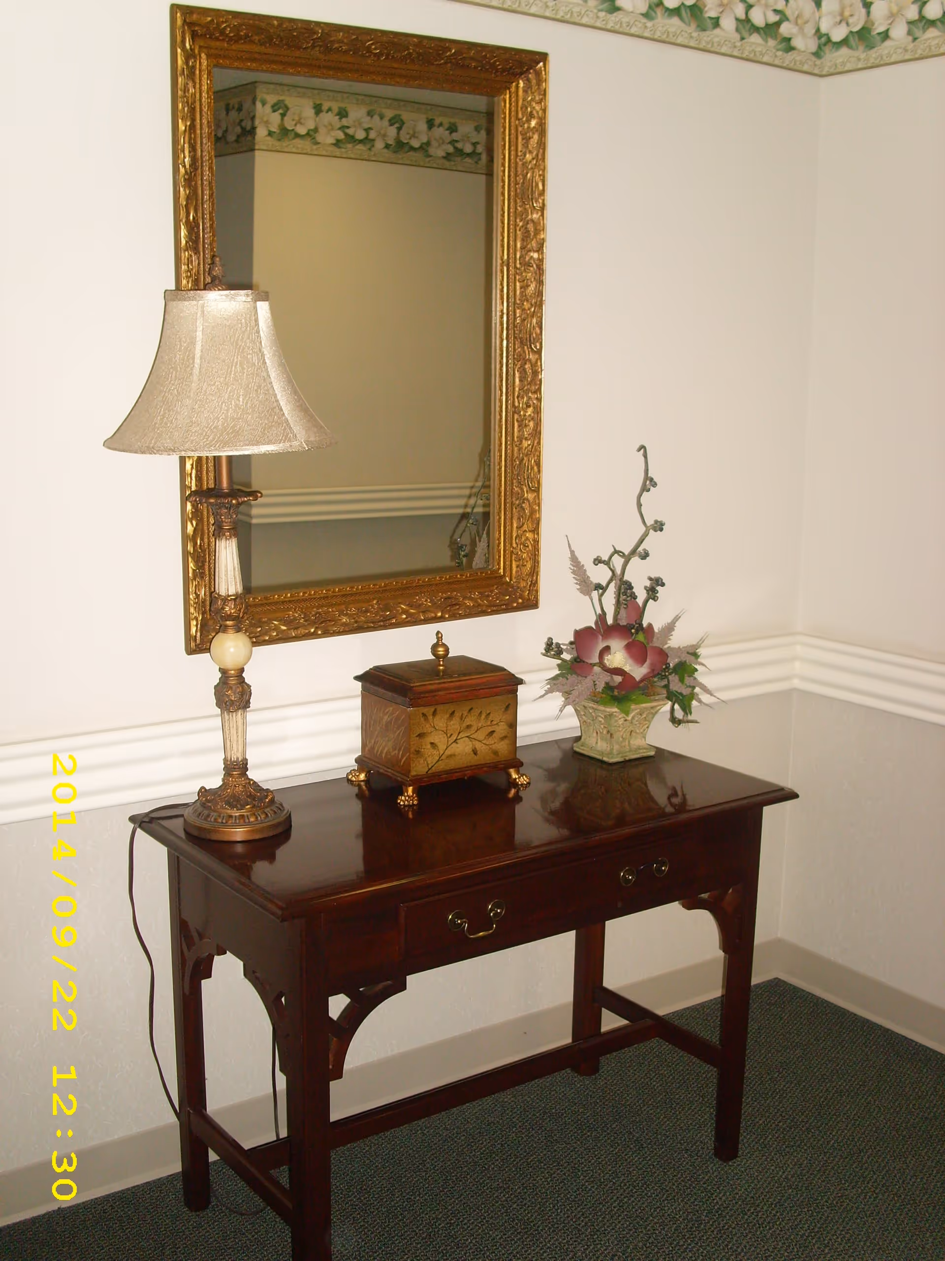 A wooden console table with two drawers against a wall, featuring a decorative table lamp, a small ornate box, and a floral arrangement in a vase. Above the table is a large, ornate gold-framed mirror. The wall has a floral wallpaper border near the ceiling and white chair rail molding.