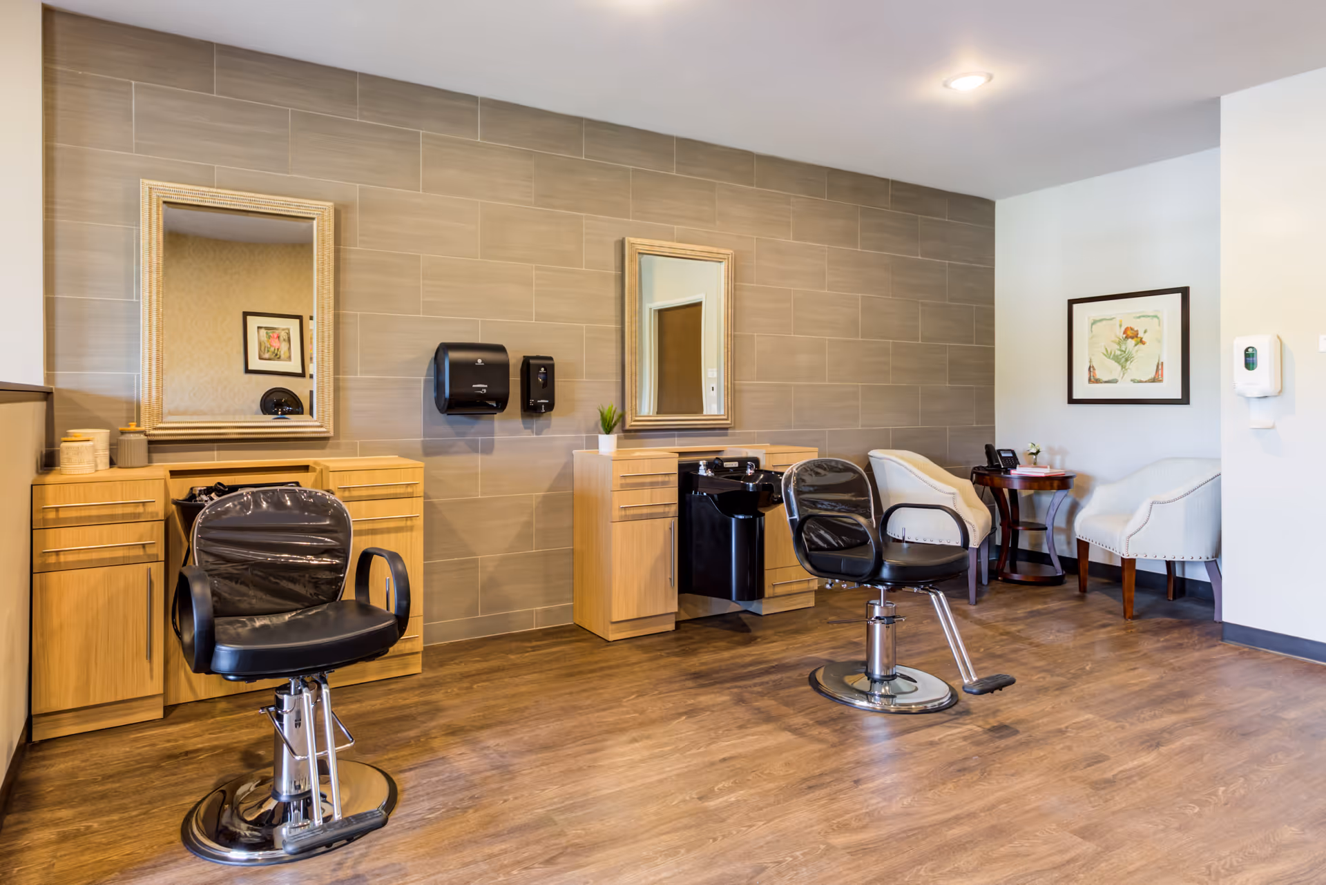 A bright interior hair salon with two styling chairs, mirrors, wooden cabinets, and a small seating area.