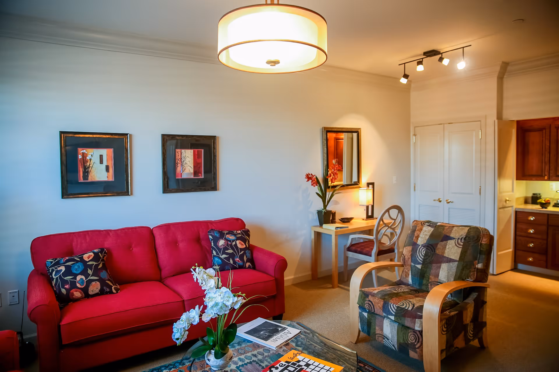 A cozy living room with a bright red sofa adorned with floral patterned cushions, a glass coffee table with a flower vase and magazines, a patterned armchair with wooden armrests, a small wooden desk with a chair, a lamp, and a mirror on the wall. The room is warmly lit with ceiling lights and has a view into a kitchen area with wooden cabinets.