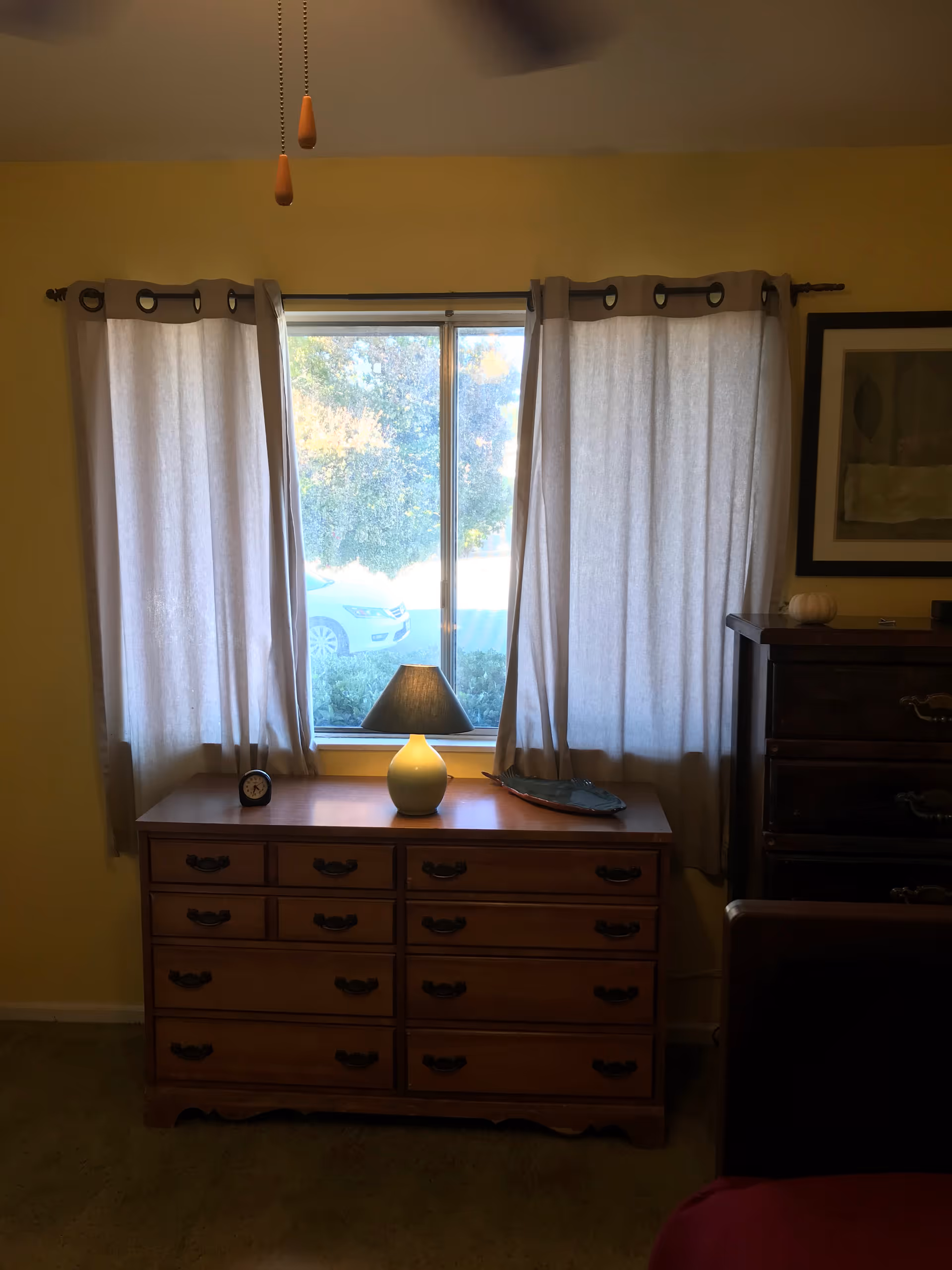A bedroom interior with a wooden dresser placed against a wall under a window with light-colored curtains. On the dresser, there is a small lamp with a dark shade, a small clock, and a decorative item. Part of a dark wooden chest of drawers and a framed picture on the wall are visible to the right.
