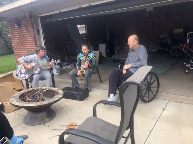 Three people sitting outside a garage. One person is playing a guitar, another is playing a saxophone, and an elderly person is sitting in a wheelchair watching them. There is a fire pit in front of them and patio chairs around.