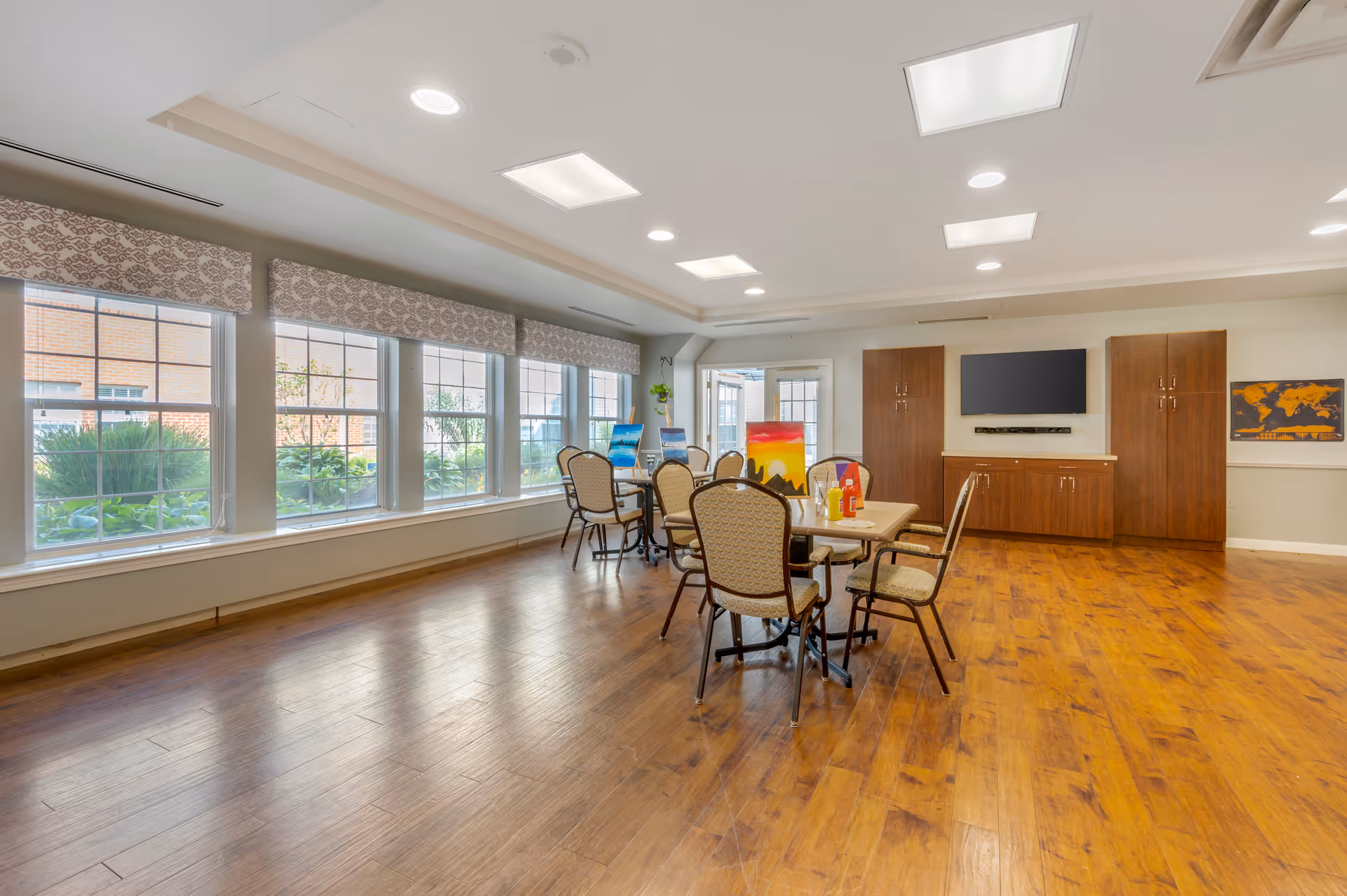 A bright and spacious common area with large windows letting in natural light. The room features wooden flooring, a table with six cushioned chairs, a wall-mounted flat screen TV, wooden cabinets, and a world map on the wall. There are some colorful paintings on easels near the windows.