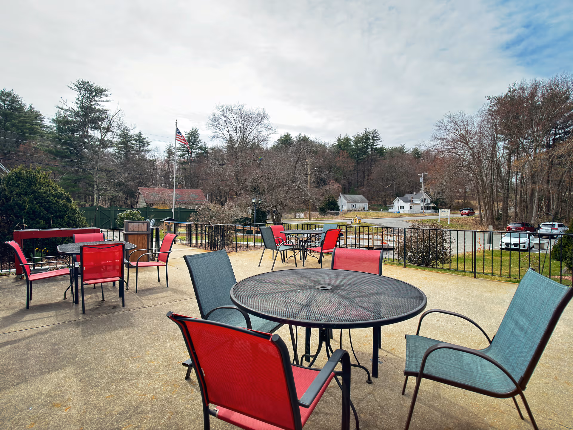 Outdoor patio area with several round metal tables and red and green mesh chairs. The patio overlooks a parking area and a road, with trees and houses in the background under a cloudy sky.
