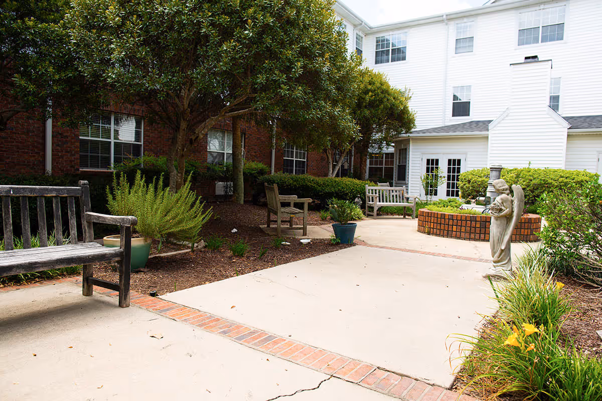 Outdoor courtyard area with wooden benches, potted plants, trees, a brick circular planter, and a small angel statue. The courtyard is surrounded by a multi-story building with white siding and brick walls.