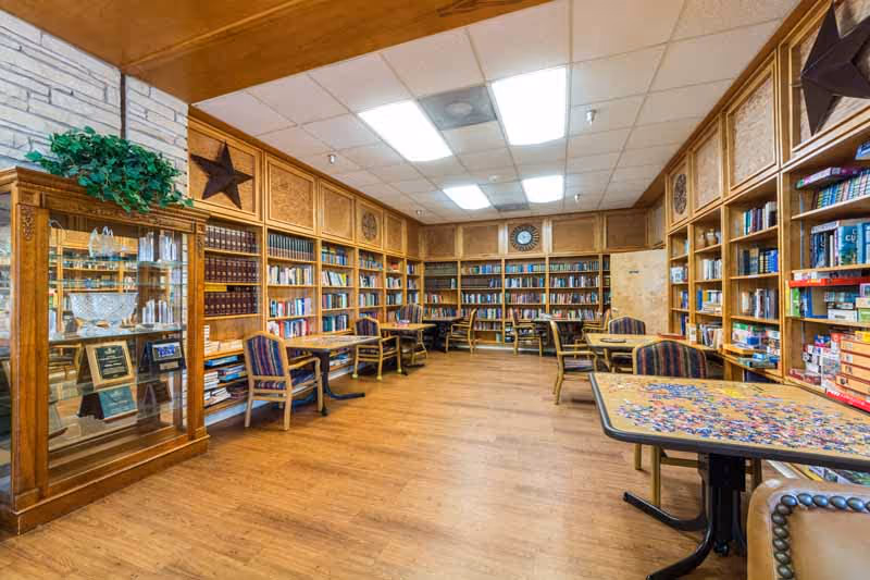 A spacious room with wooden floors and walls lined with bookshelves filled with books and board games. Several tables and chairs are arranged around the room, including one table with a partially completed jigsaw puzzle. A glass display cabinet with trophies and plaques is visible on the left side. The ceiling has fluorescent lighting and a clock is mounted on the far wall.