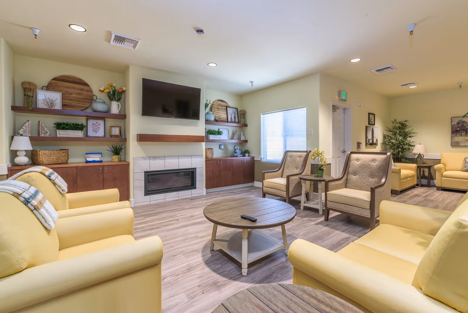 A cozy living room area in a senior living facility with yellow armchairs, beige cushioned chairs, a round wooden coffee table with a remote control, built-in shelves decorated with plants, pictures, and decorative items, a wall-mounted flat-screen TV above a modern fireplace, and a window letting in natural light.