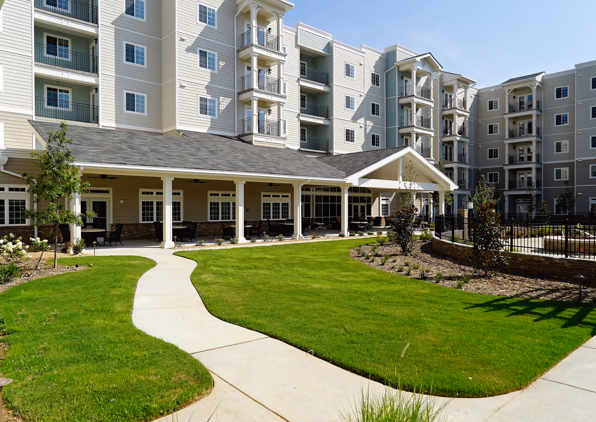 Outdoor view of Towne Club Windermere showing a well-maintained green lawn with a winding concrete pathway leading to a covered patio area with tables and chairs. The multi-story building with balconies and numerous windows surrounds the courtyard under a clear blue sky.