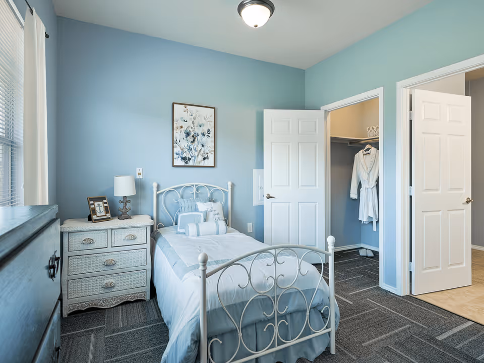 A cozy bedroom with light blue walls, a single white metal bed with blue and white bedding, a wicker nightstand with a lamp and picture frame, a window with white curtains, and an open closet door showing a white robe hanging inside.