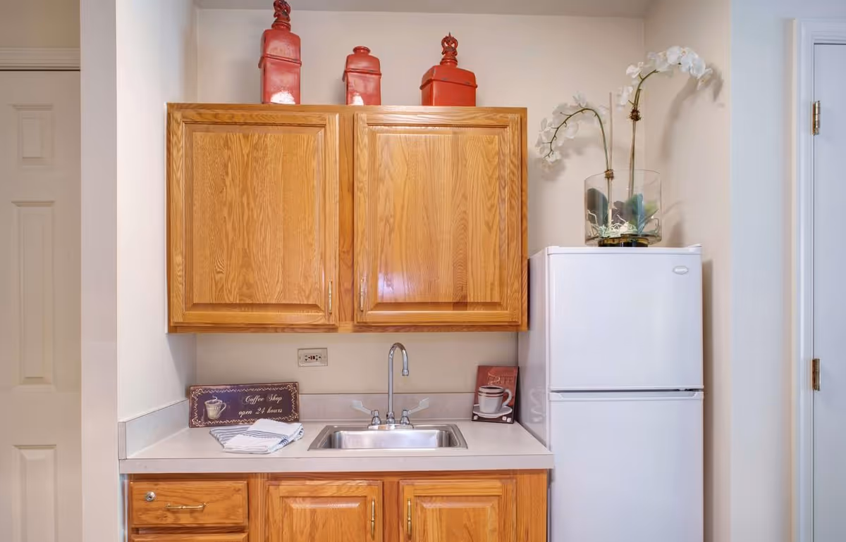 Small kitchenette with oak cabinets, a stainless sink, white refrigerator and decorative items on the counter and atop the cabinets.