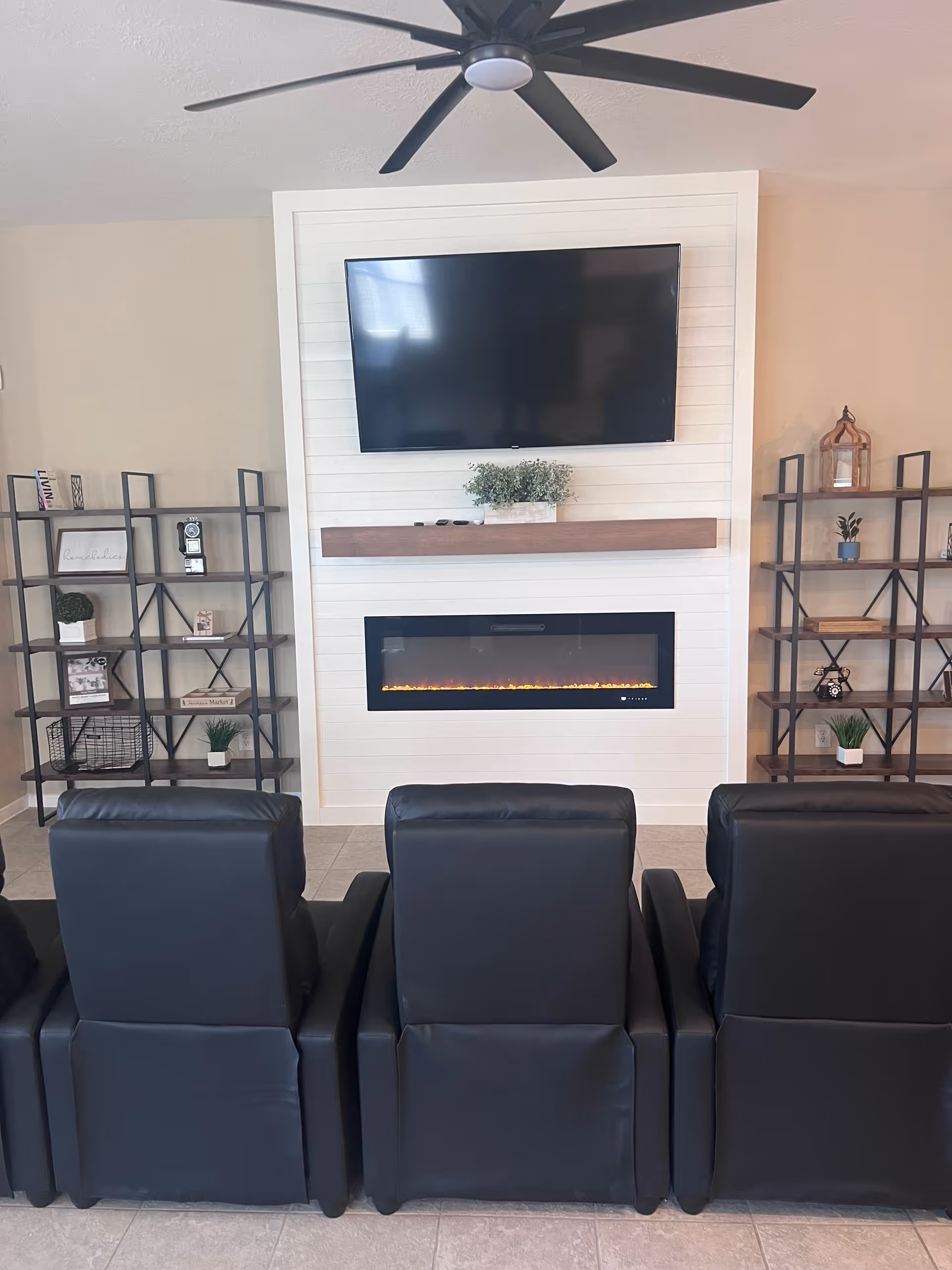 A cozy living room area with three black leather recliners facing a wall-mounted flat-screen TV above a modern electric fireplace. On either side of the fireplace are black metal shelving units with decorative items and plants. A large black ceiling fan is visible above.