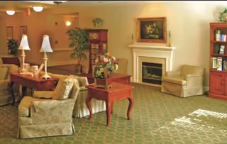 Well-lit common room with sofas, armchairs, side tables, lamps, a fireplace, and a bookshelf.