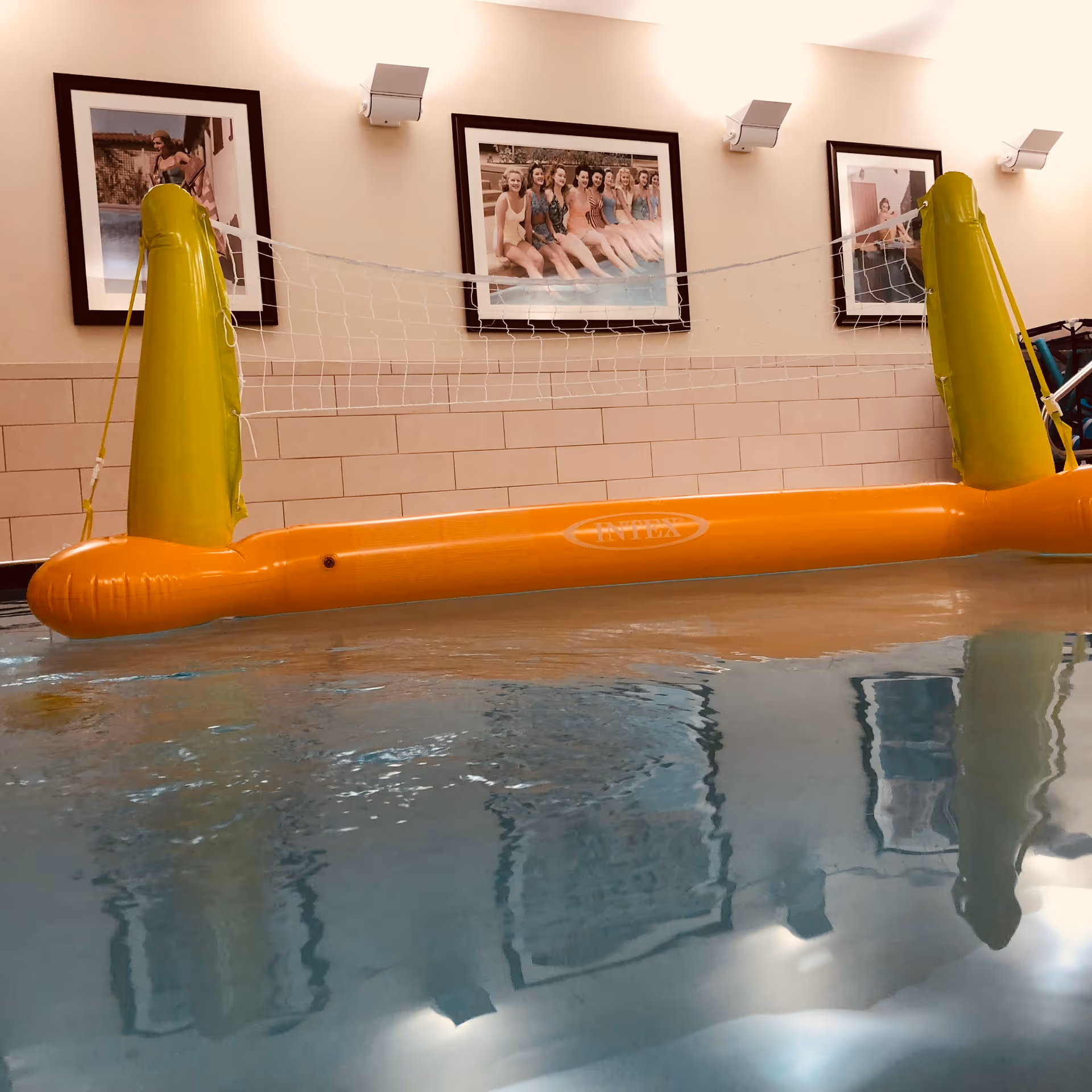 Indoor swimming pool with an inflatable volleyball net set up across the water. The wall behind the pool has three framed vintage-style photos of women in swimsuits sitting by the pool. The pool water reflects the images and lights on the wall.