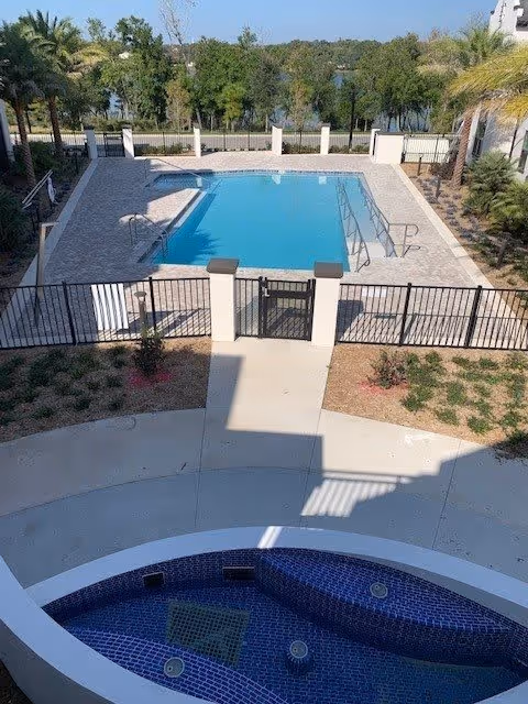 View of a fenced outdoor swimming pool with clear blue water, surrounded by a paved deck and palm trees. In the foreground, there is a curved blue-tiled hot tub or spa area. Beyond the pool area, there are trees and a body of water visible in the background.