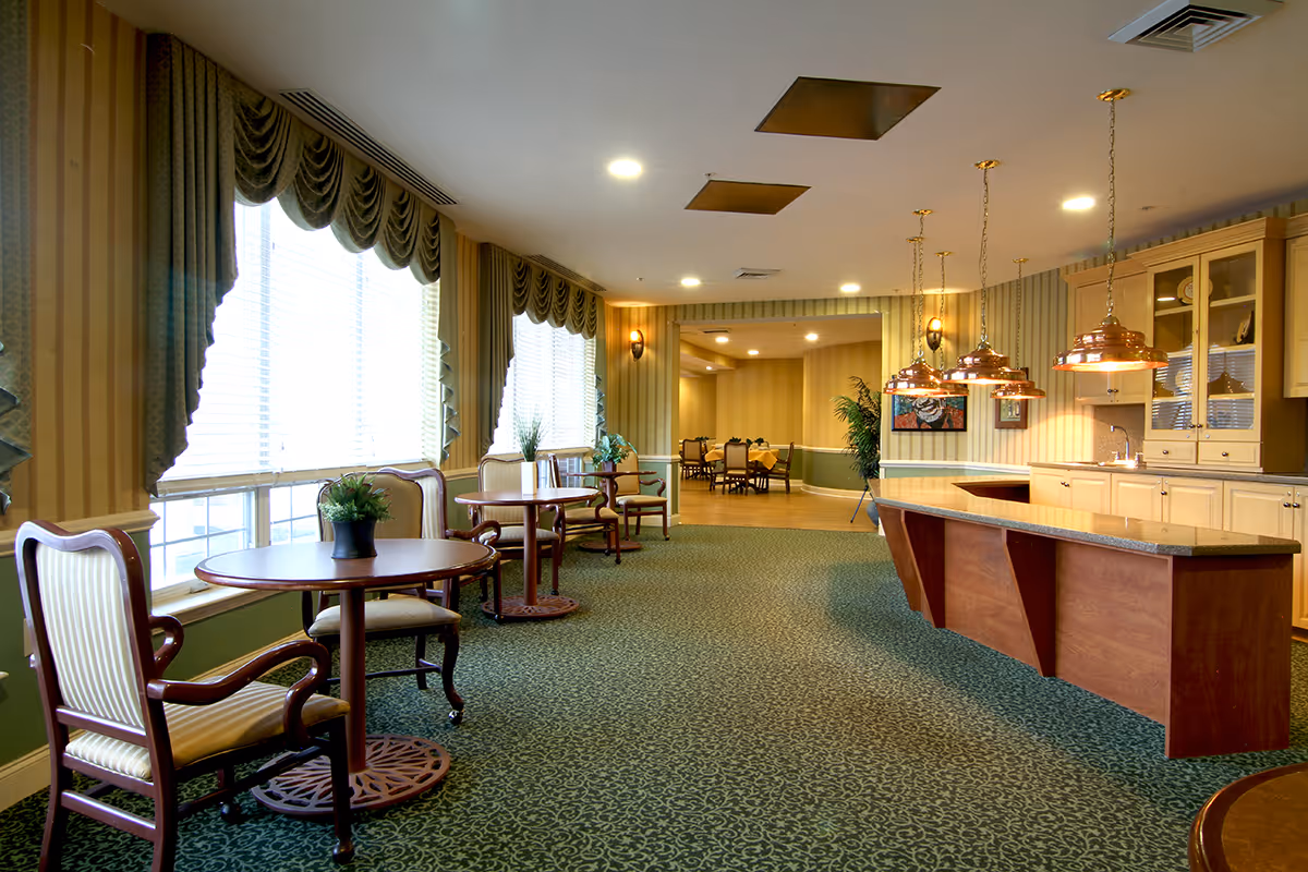 A spacious, well-lit interior room with large windows covered by green drapes. The room features round wooden tables with cushioned chairs, a long wooden counter with hanging copper pendant lights, and a carpeted floor with a green patterned design. In the background, there is another dining area with more tables and chairs, and decorative plants and artwork on the walls.