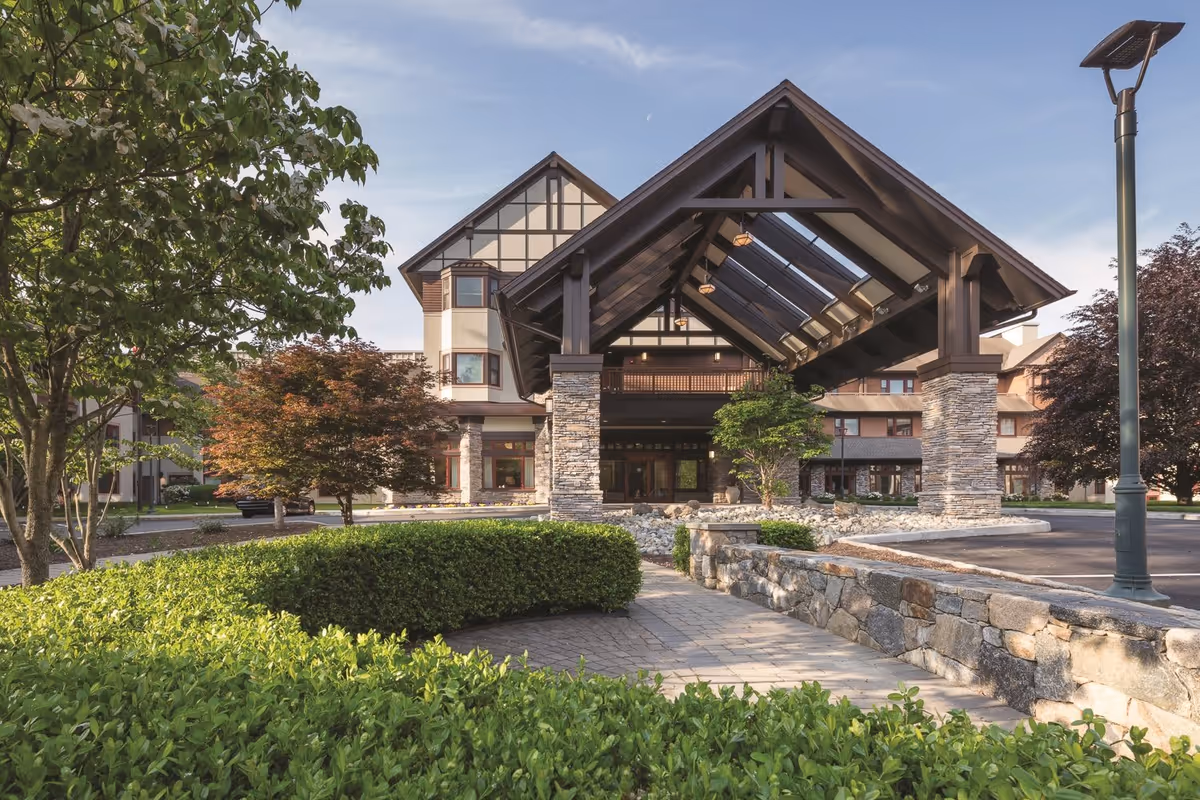 Front exterior view of Edgehill Senior Living Community Stamford showing a large covered entrance with stone pillars, surrounded by well-maintained greenery and trees under a clear sky.