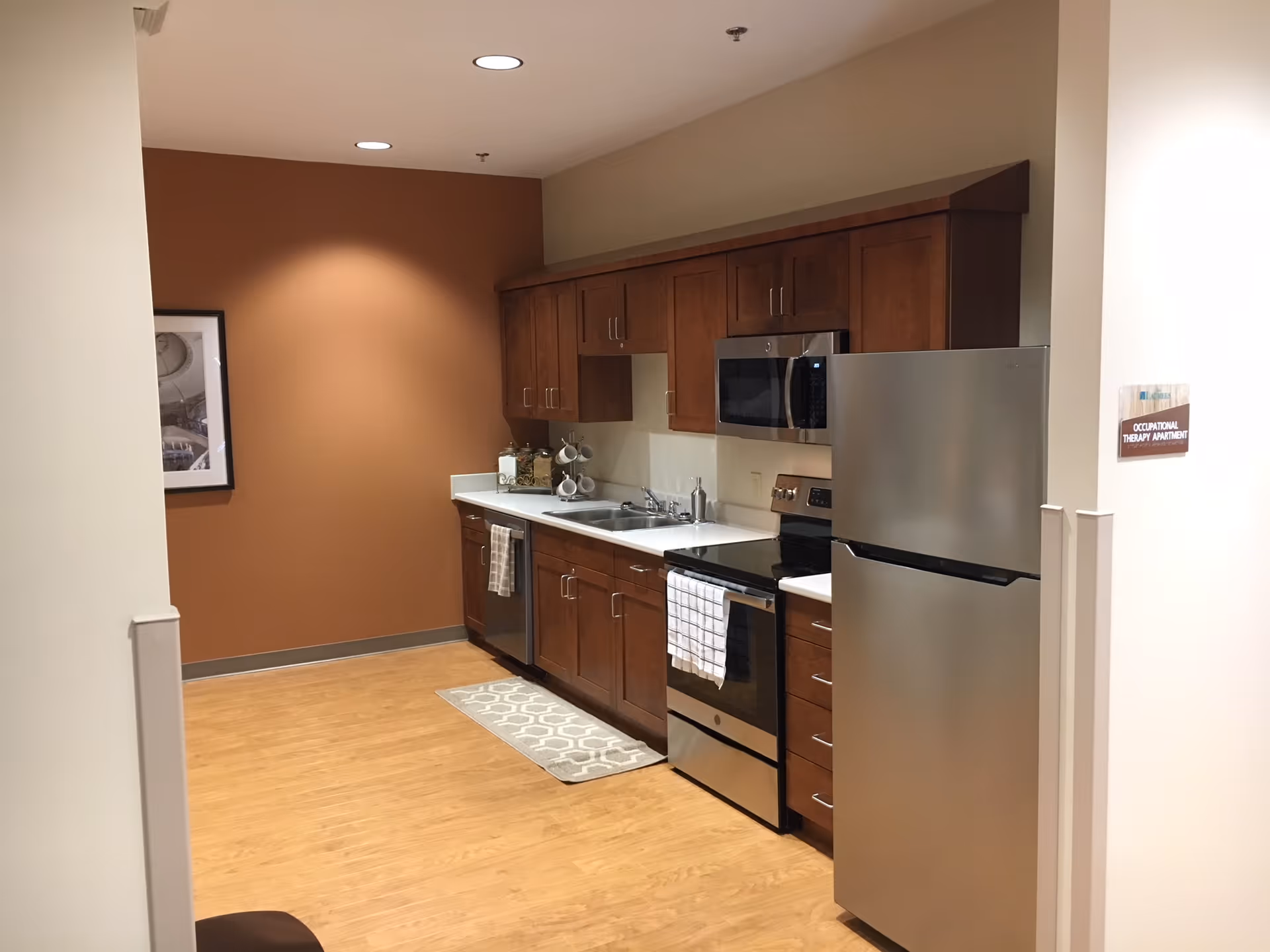 Modern kitchen area with wooden cabinets, stainless steel refrigerator, oven, microwave, and dishwasher. The kitchen has a white countertop, a sink, and a patterned rug on the wooden floor. The walls are painted in beige and brown tones, and there is a framed picture on the left wall.