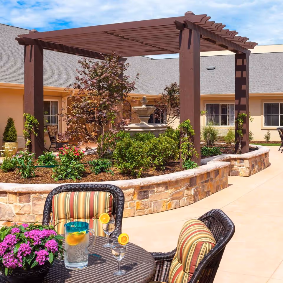 Sunlit courtyard with a wooden pergola over a planted bed and fountain, and a patio table with striped chairs and a pitcher of lemon water.