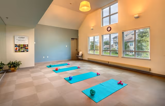 A bright exercise room with four turquoise yoga mats laid out on a checkered floor, each mat accompanied by small dumbbells and exercise balls. The room has large windows letting in natural light, a high ceiling with a hanging light fixture, a clock on the wall, and a chair in the corner. There is a plant and a framed poster on the left wall.