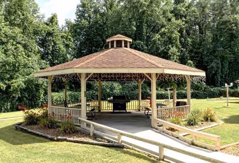 Wooden gazebo with benches sits on a grassy lawn, surrounded by trees.